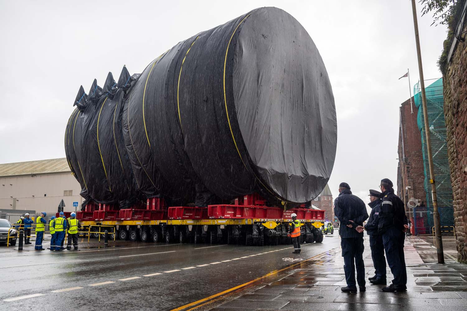 Building-sized section of submarine covered in black tarpaulin sits on a many-wheeled flatbed truck that is manoeuvring onto a street. Figures in high vis vests stand near the vehicle, and three figures in naval attire observe them from the pavement. It is raining and a police car is partially visible in the background, blocking the traffic.