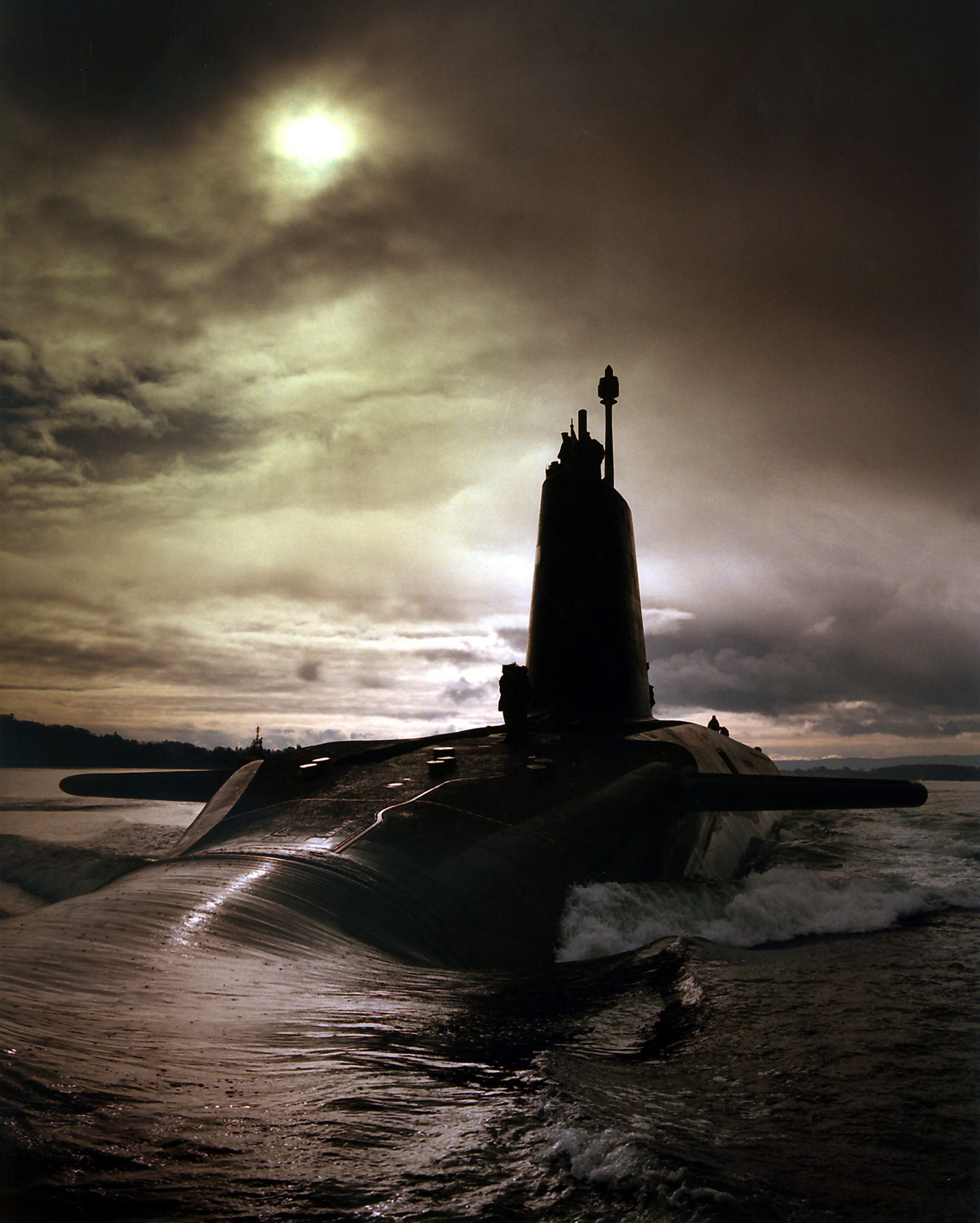 A submarine sails away from the viewer, it's fin silhouetted against a cloudy sky with the sun just distinguishable through the grey.