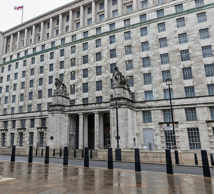 The facade of an archetypal Whitehall building, constructed in Portland stone, with barred windows, two statues in either side of the entrance and a flag at the top. Viewed from across the road on a grey, raining day, the building looks both imposing and dreary.