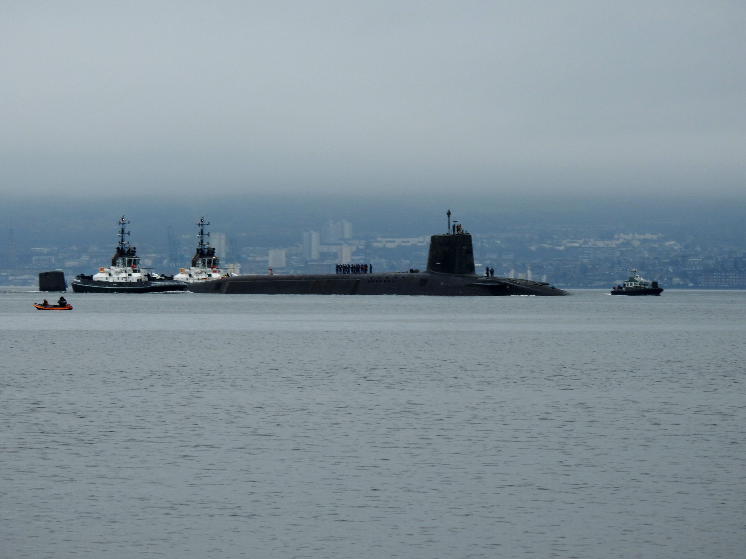 Two tug boats help to push a long dark submarine through the water, under a low grey clouds. Crew members are lined up on the submarine hull