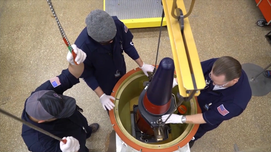 Three workers are standing around a canister, which we see from above. A crane in the foreground is pulling a warhead out of the canister, under the control of one of the workers