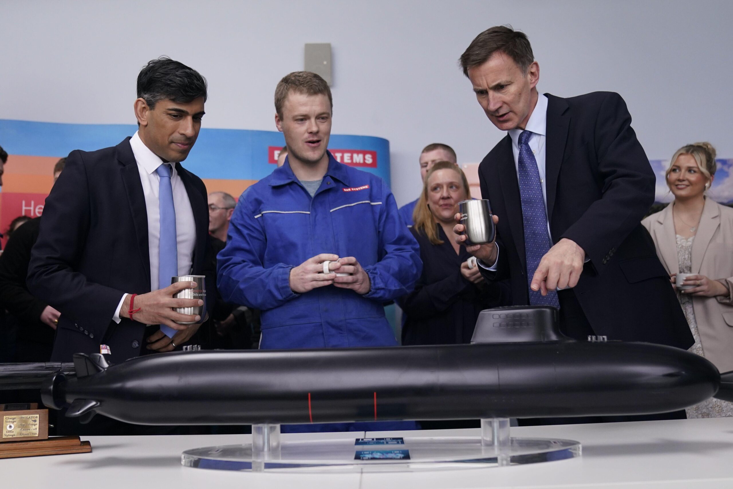 Rishi Sunak and Jeremy Hunt stand behind a mockup model of an SSN-AUKUS submarine with a worker in blue overalls standing between them.